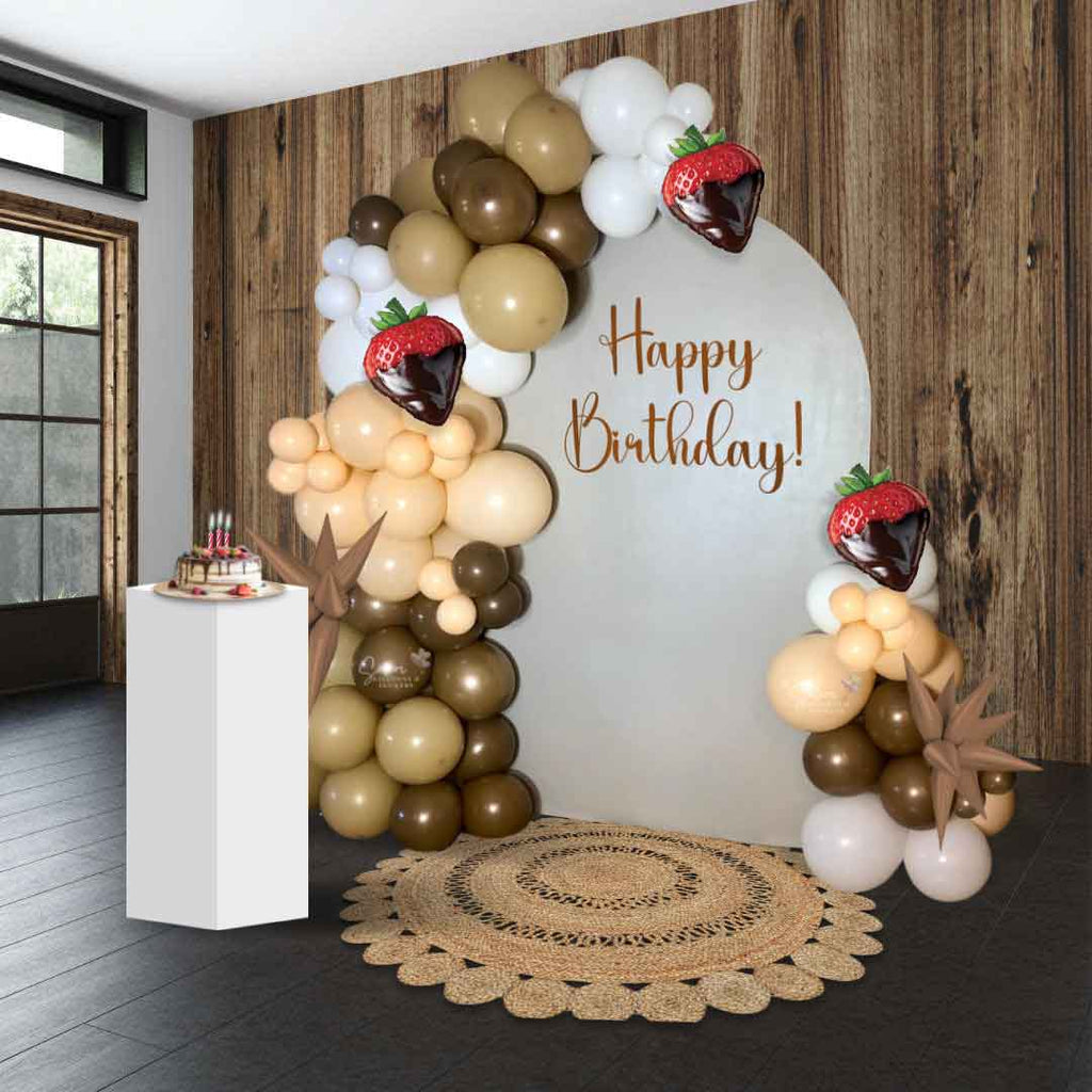 A wooden wall and brown floor room with a  balloon garland decorating a single panel with a Happy Birthday sig. Strawberries and Chocolate theme in a color palette of Chocolate Brown, caramel, white, and red. Oregon Balloon Decor