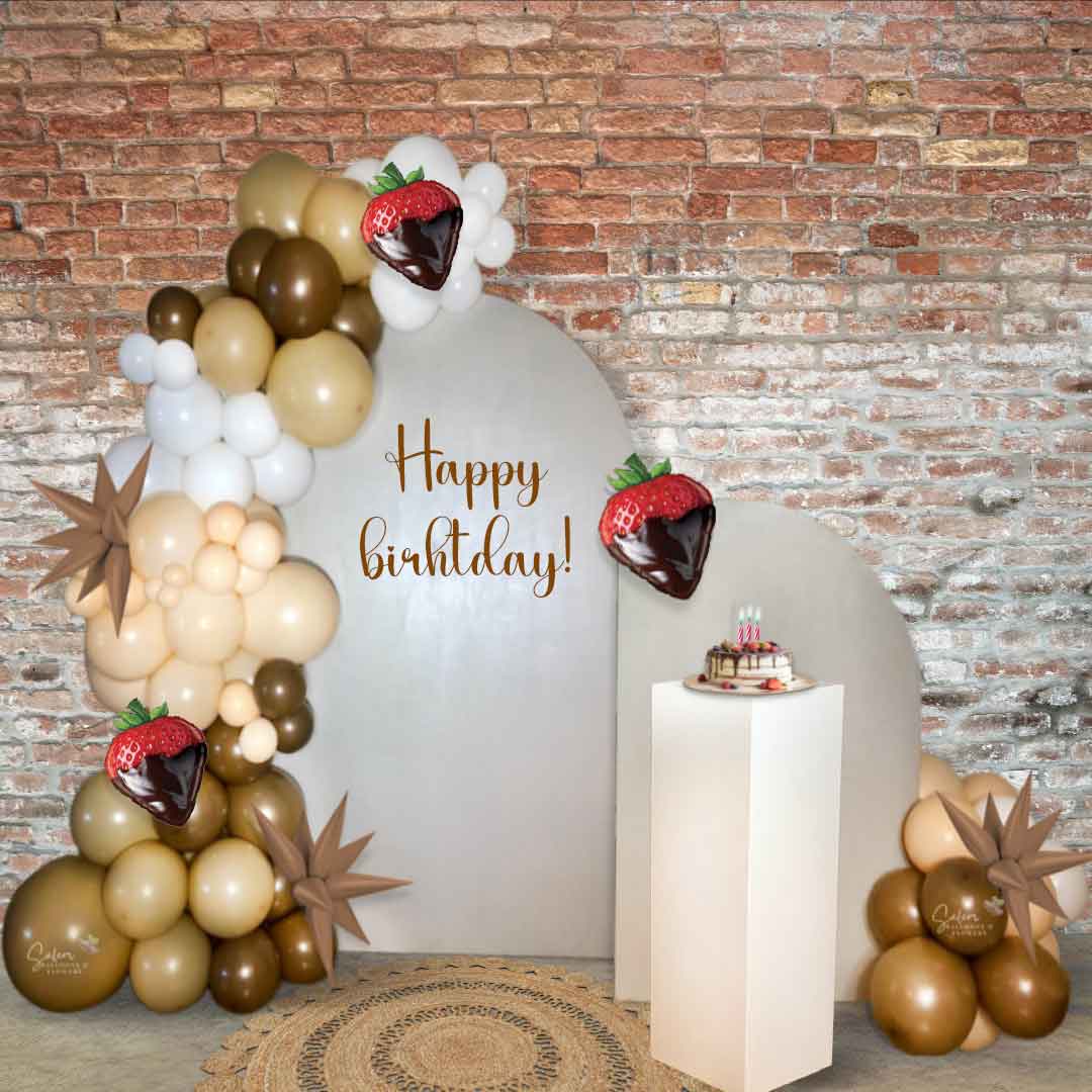A 2 panel-2 garland Strawberries and Chocolate themed Balloon Decoration with a white cake stand and a round jute rug in front of a brick wall. . Oregon Balloon Decor.