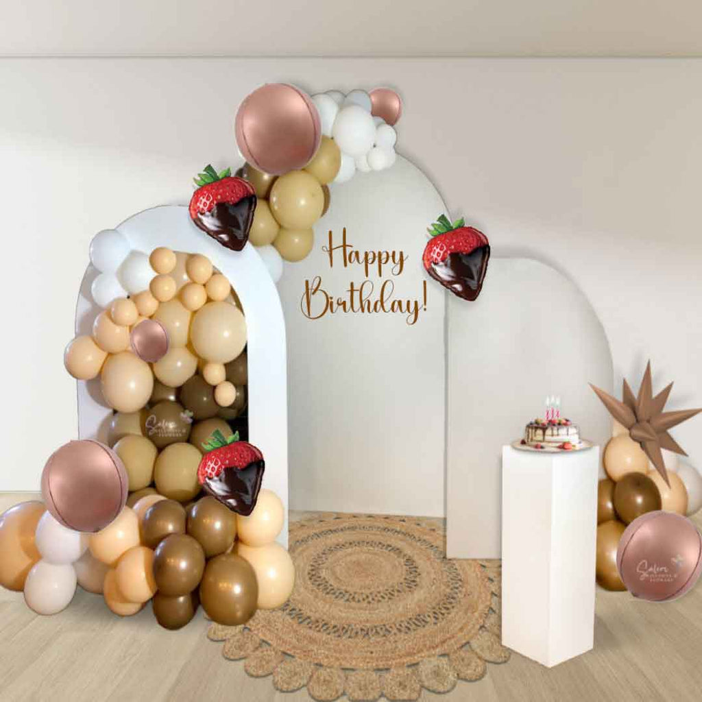 A White room decorated with a 3 panel 2 garland strawberries and chocolate themed balloon decorations, with a cake stand and a round jute rug. Oregon Balloon decor.