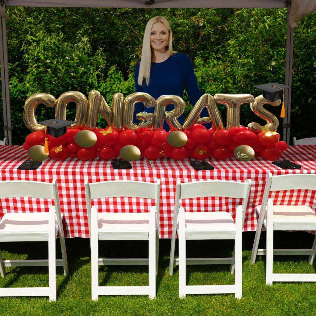 A lady standing behind a table with a Balloon table runner spelling CONGRATS with gold balloon letters on a balloon garland. Salem Oregon Balloon decor delivery