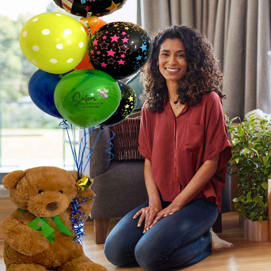 A woman kneeling on the floor next to a Large teddy bear holding a set of helium-filled balloons with a 