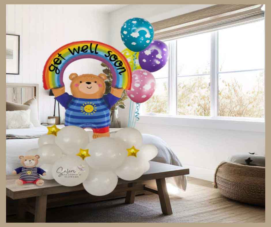 A colorful teddy bear balloon arrangement with rainbow balloons and stars, standing on a bench next to a teddy bear plush in front of a bed. Oregon Balloon Delivery.