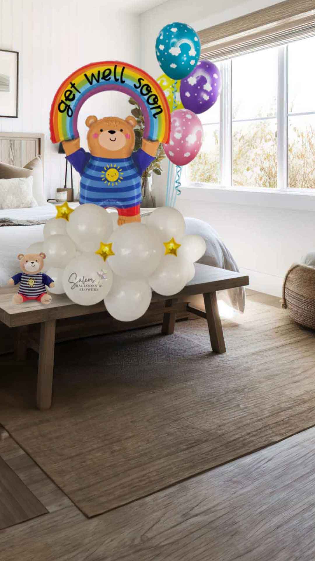 A colorful teddy bear balloon arrangement with rainbow balloons and stars, standing on a bench next to a teddy bear plush in front of a bed. Oregon Balloon Delivery.