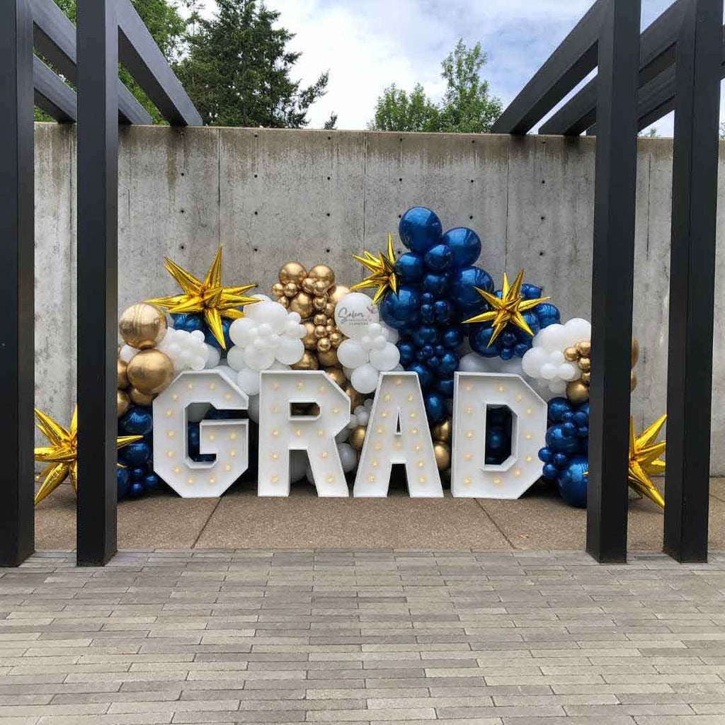 Grad Balloon decor display. Featuring a GRAD light up marquee letters nested in a zaphire blue, gold and white balloon wall. Salem Oregon Balloon decor and marquee letters rental