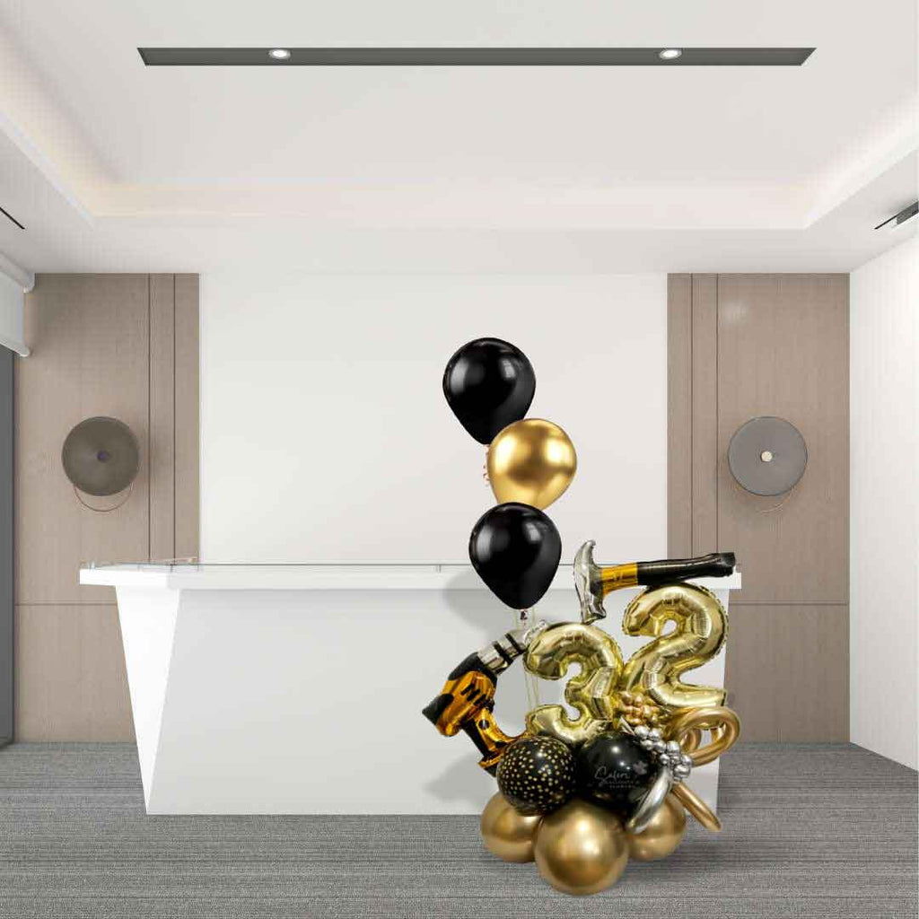 Standing on the floor in front of a desk it's a Number Balloon Arrangement, featuring a balloon numbers, Balloon hammer and a drill, decorated with curly balloons and  a set of helium balloons in gold, black and some touches of silver. Oregon Balloon Delivery.