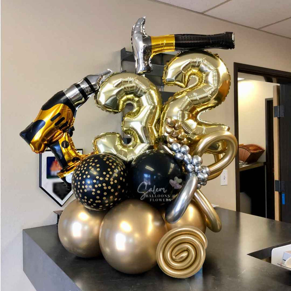 Standing on a desk is a Number Balloon Arrangement, featuring a balloon hammer and a drill, decorated with curly balloons in gold, black and some touches of silver. Oregon Balloon Delivery