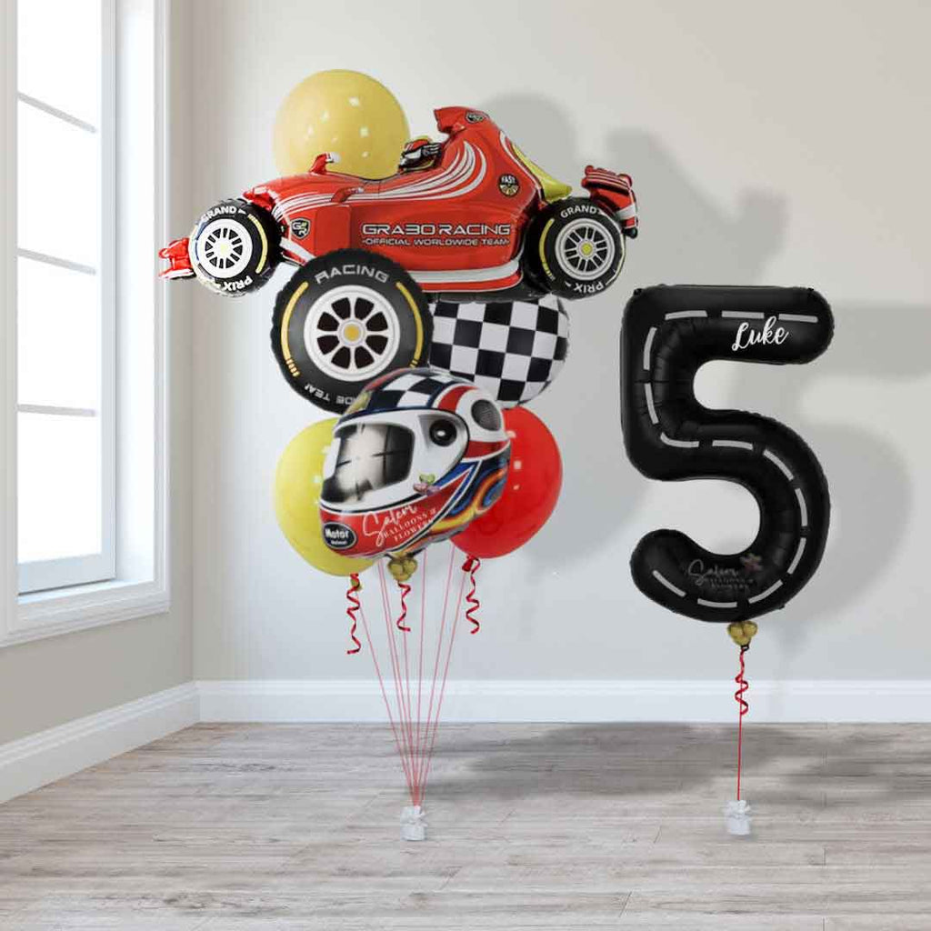 A tall helium Balloon bouquet, featuring a red sports car shaped balloon, a helmet shaped balloon, a race car wheel balloon and Latex balloons in Red, black, yellow and touches of blue next to a black number balloon 5 with a race track design in front of a window. Oregon balloon delivery.