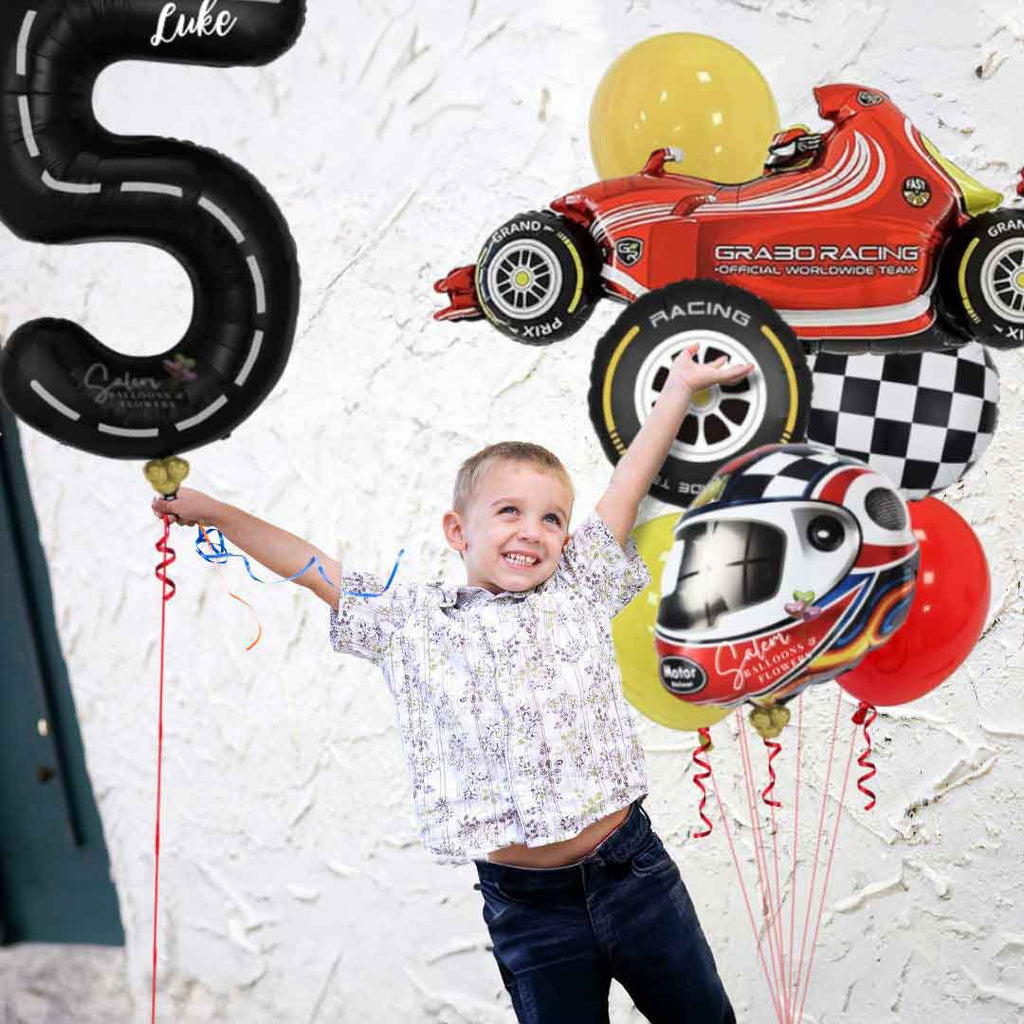 A boy holding a tall helium Balloon bouquet, featuring a red sports car shaped balloon, a helmet shaped balloon, a race car wheel balloon and Latex balloons in Red, black, yellow and touches of blue, and a black number balloon 5 with a race track design. Salem Oregon birthday balloon delivery.