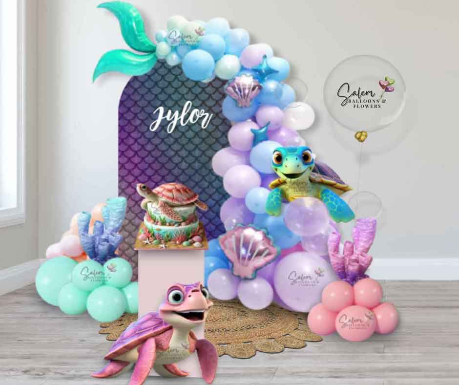 Mermaid and turtles-themed balloon decoration by Salem Balloons and Flowers. Features a tall mermaid tail balloon garland in purple, blue, and pink shades, attached to a mermaid scale Chiara panel. Decorated with coral accents and adorable turtle cutouts. Perfect for underwater and mermaid-themed celebrations, available in Salem, Oregon, and nearby cities.