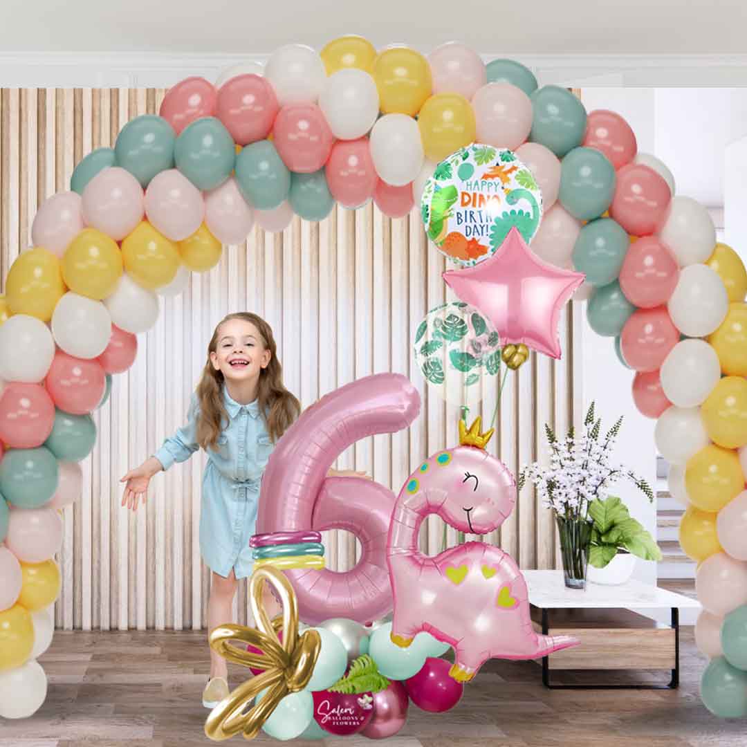 a little girl standing with a balloon decoration of 1 balloon arch and a Pink dinosaur themed number balloon marquee. Featuring a large pink dinosaur wearing a golden crown, decorated with helium balloons. Balloons salem oregon. Balloon decor.