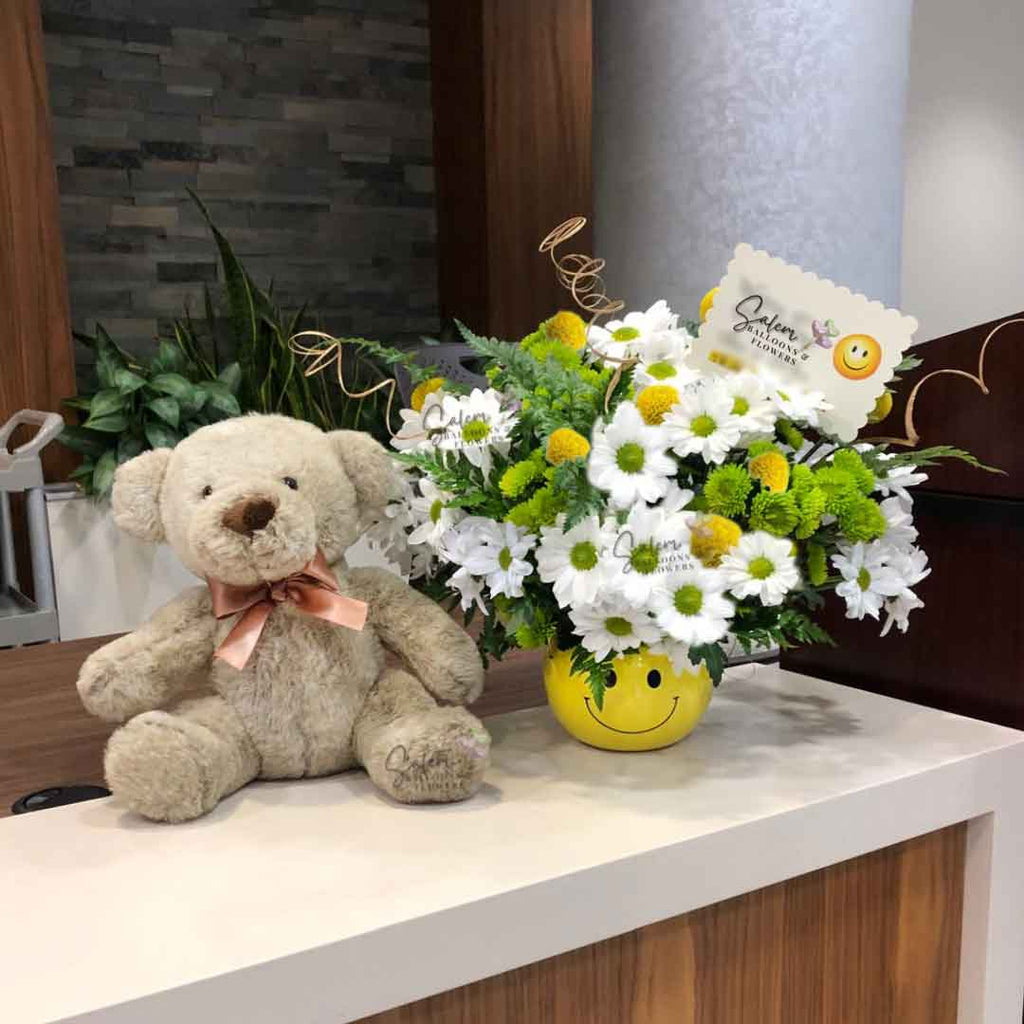 Yellow Smiley face vase with white daisies, green mooms, curly willow and yellow pum-pums. Next to a teddy bear plush. Flower delivery. Salem Oregon.