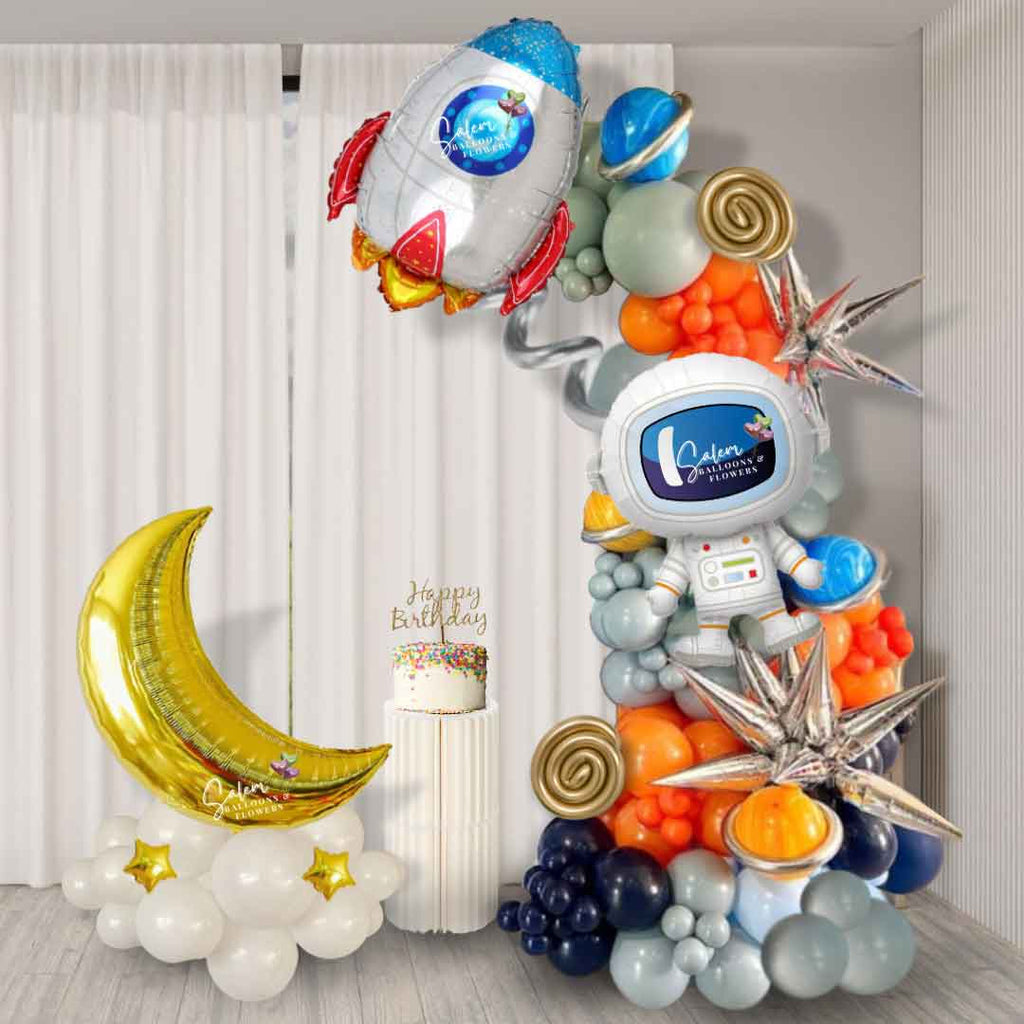 Space-themed balloon decor in silver, orange, and blue. Showcasing an 8-foot organic garland adorned with an astronaut foil balloon, star and planet accents, and a Moon satellite balloon arrangement. Perfect for events in Salem, Oregon, and surrounding areas.