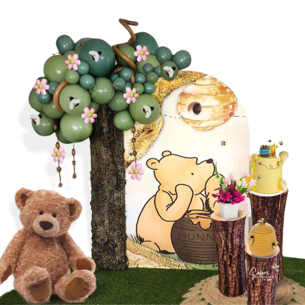 A Winnie the Pooh Balloon decoration, featuring a balloon tree next to a Winnie the Pooh backdrop, 3 cake stands and a teddy bear, all on a grass rug. Oregon Balloon Decor.
