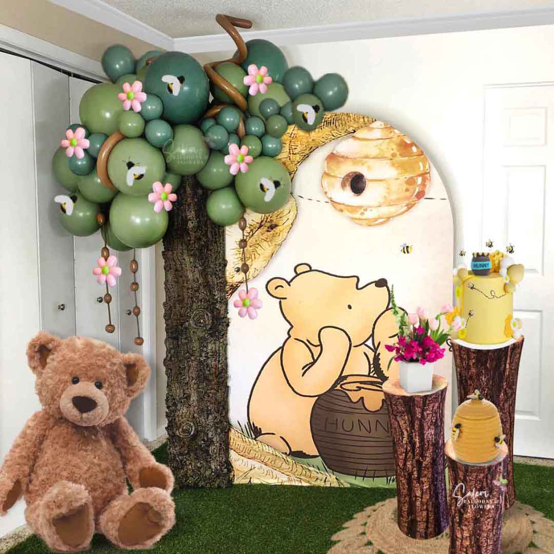 A Winnie the Pooh Balloon decoration, featuring a balloon tree next to a Winnie the Pooh backdrop, 3 cake stands and a teddy bear, all on a grass rug. Oregon Balloon Decor.