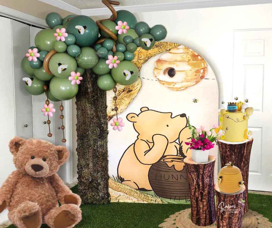 Winnie the Pooh themed balloon decoration, with balloon tree, Large teddy bear, cake stands with cake and flowers, and backdrop in a yellow, green and light blue palette. Oregon Balloon Decor. 