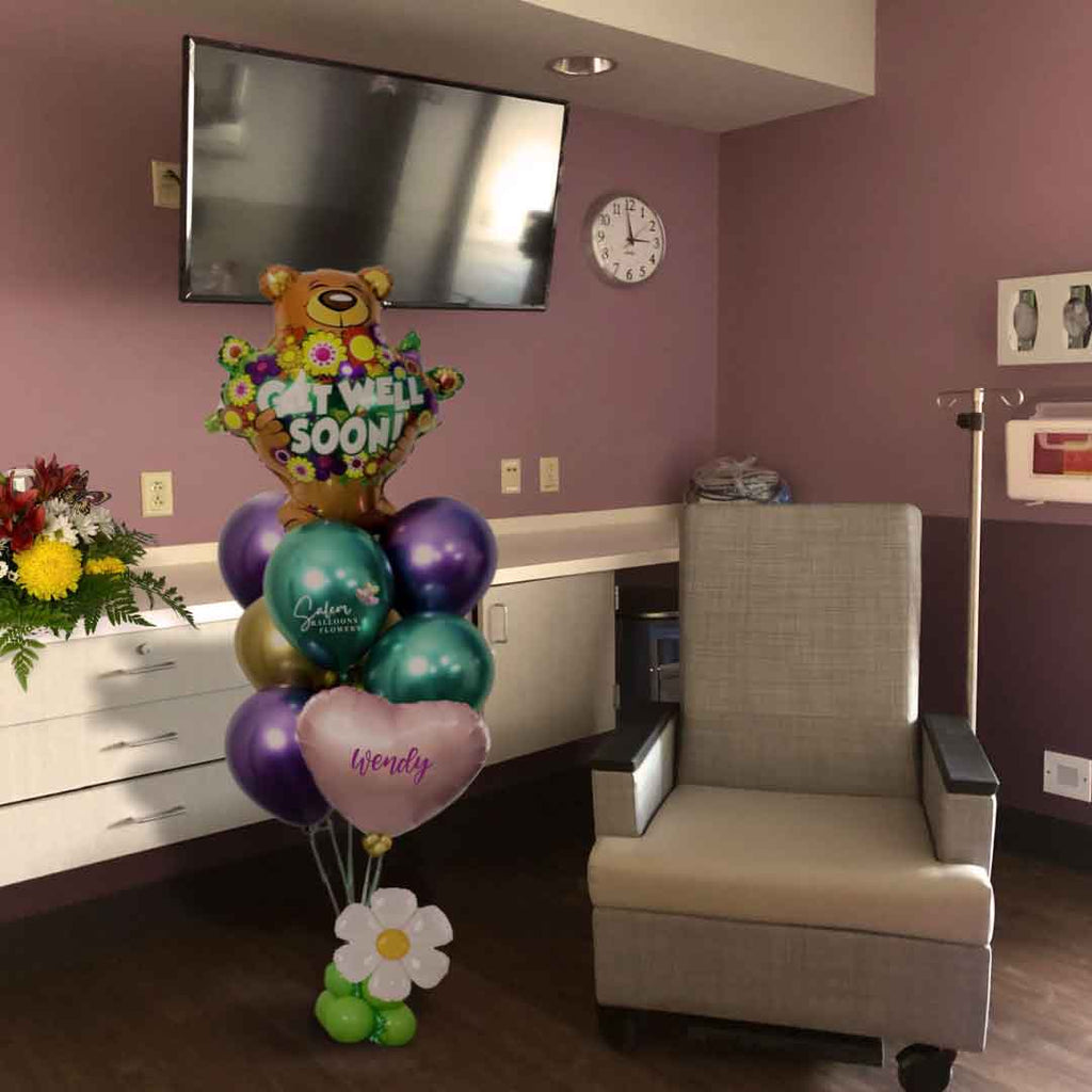 A hospital room with flowers and a Get Well helium balloon bouquet. Featuring a large teddy bear balloon holding flowers with a 