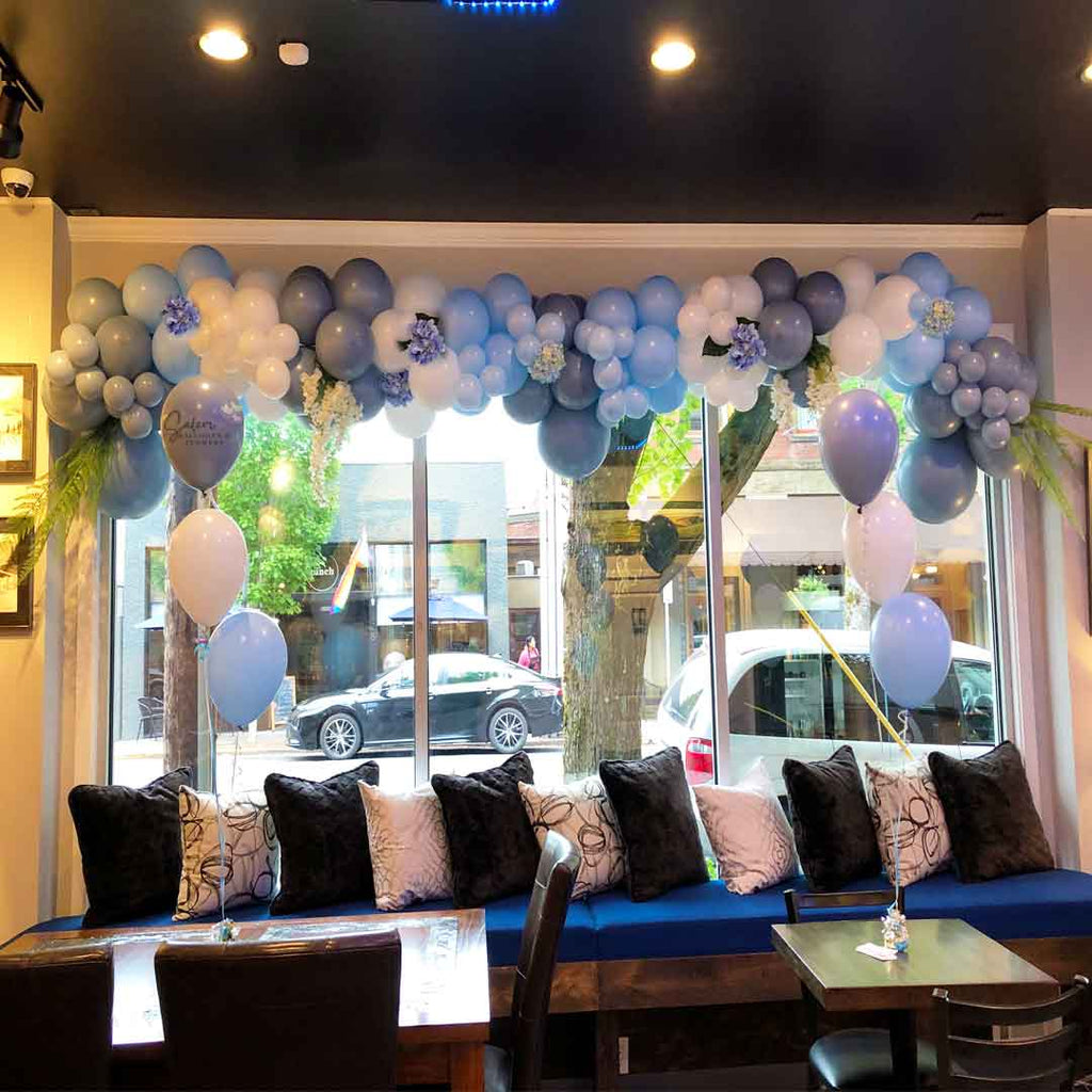 12 ft Organic balloon garland in different shades of blue and embellished with blue hydrangeas and greenery decorating a wall to wall window in a restaurant. Oregon Balloon Decor