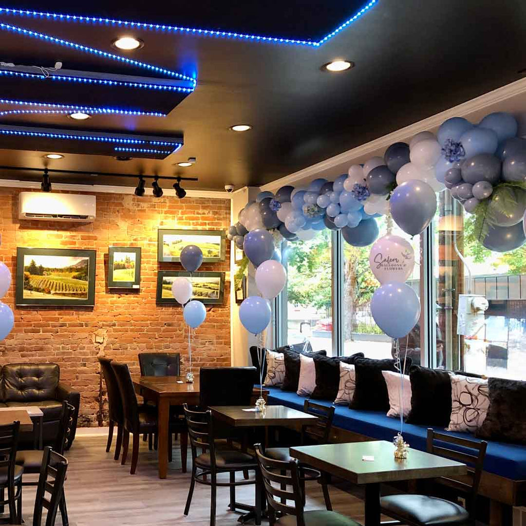 12 ft Organic balloon garland in different shades of blue and embellished with blue hydrangeas and greenery decorating a wall to wall window in a restaurant. Oregon Balloon Decor